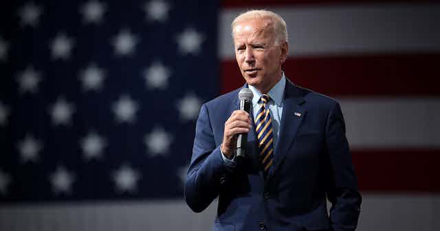 President Biden speaking with a microphone in front of an American flag.
