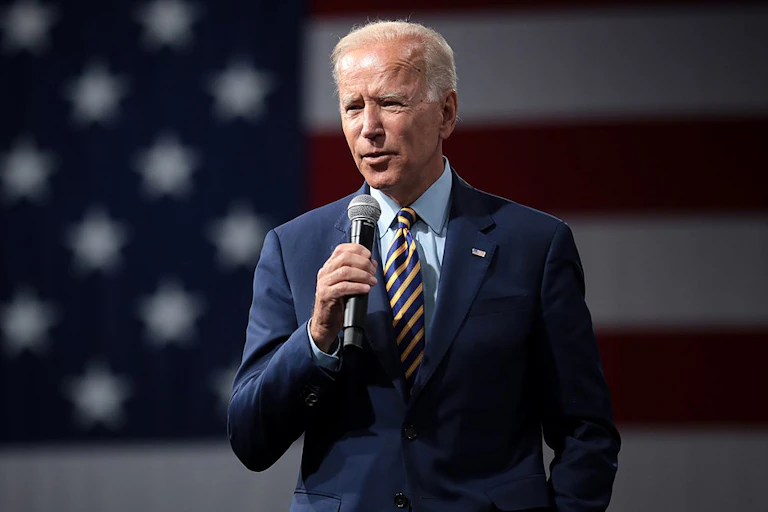 President Biden speaking with a microphone in front of an American flag.