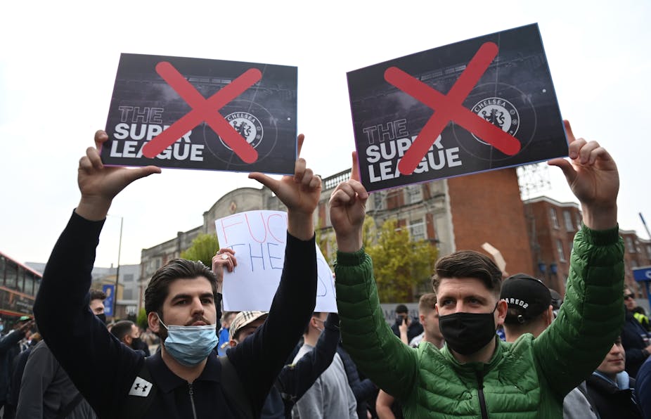 Two men wearing face masks in protest crowd hold up European Super Leaue posters with red crosses over both of them