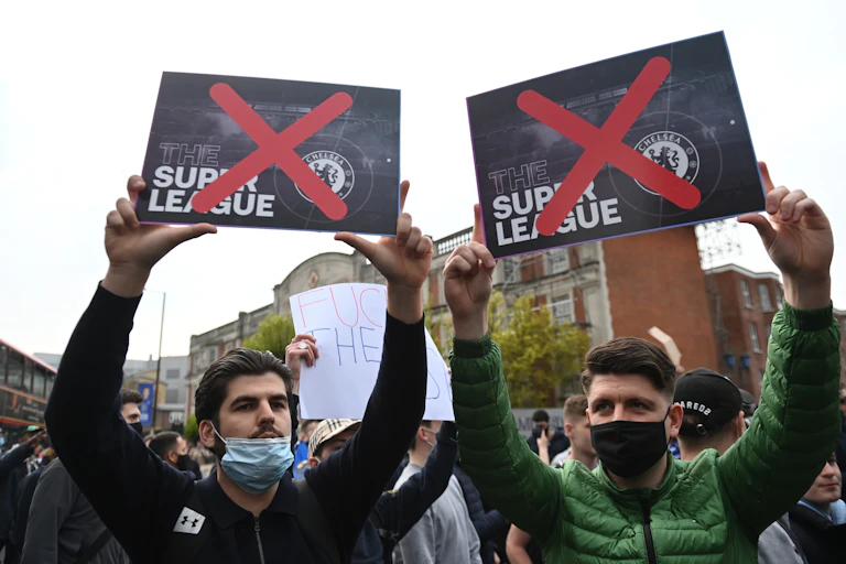 Two men wearing face masks in protest crowd hold up European Super Leaue posters with red crosses over both of them