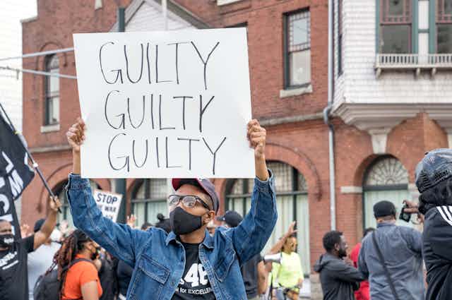 A person in a crowd holding a sign that says, "GUILTY GUILTY GUILTY"