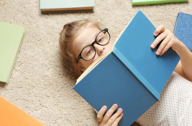 Una niña con gafas leyendo tumbada un libro de tapas azules.
