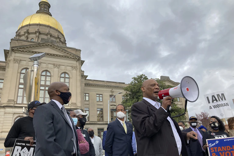 A man surrounded by other protestors with signs holds a megaphone in front of the Georgia Capitol building, with its golden dome in the distance