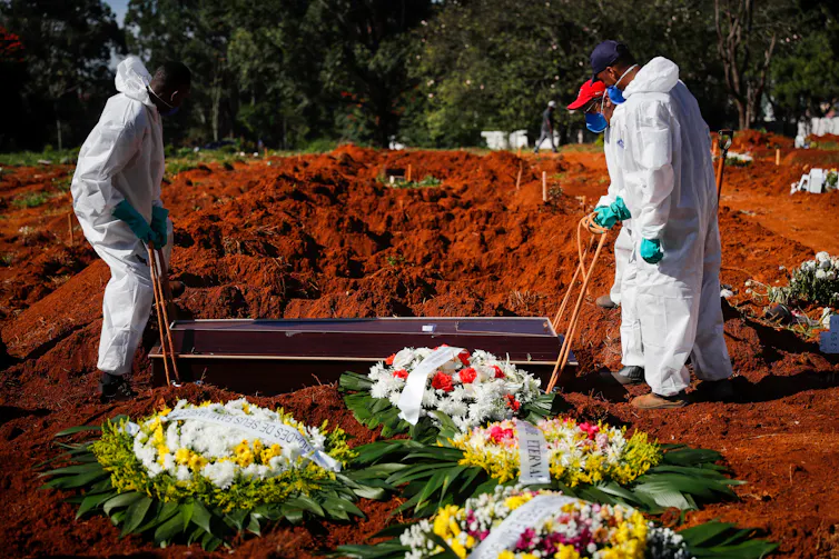 Two people in protective suits, masks and gloves lower a coffin into a grave.