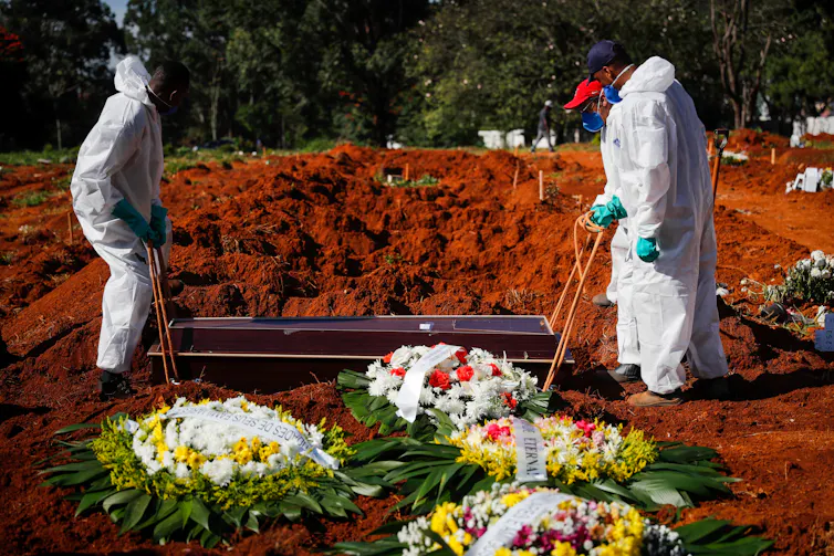 Two people in protective suits, masks and gloves lower a coffin into a grave.