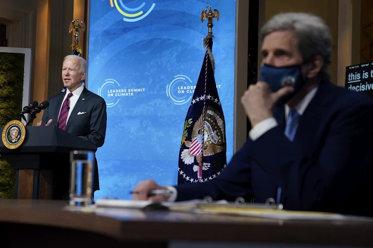 Biden speaking to the camera and Kerry, wearing a face mask, sitting to the side.