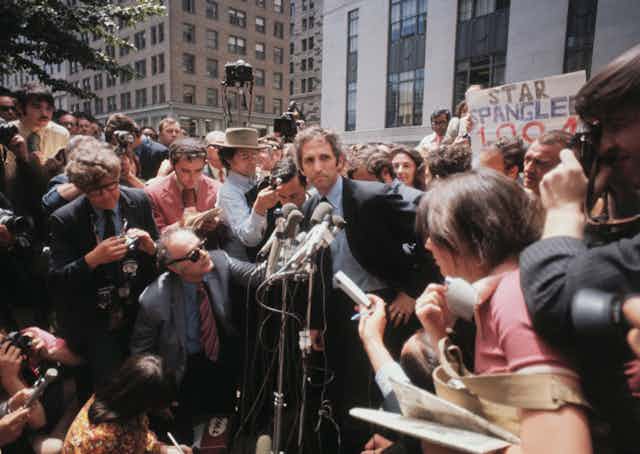 Ellsberg stands in front of microphones surrounded by reporters and photographers.