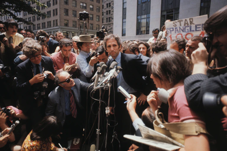 Ellsberg stands in front of microphones surrounded by reporters and photographers.