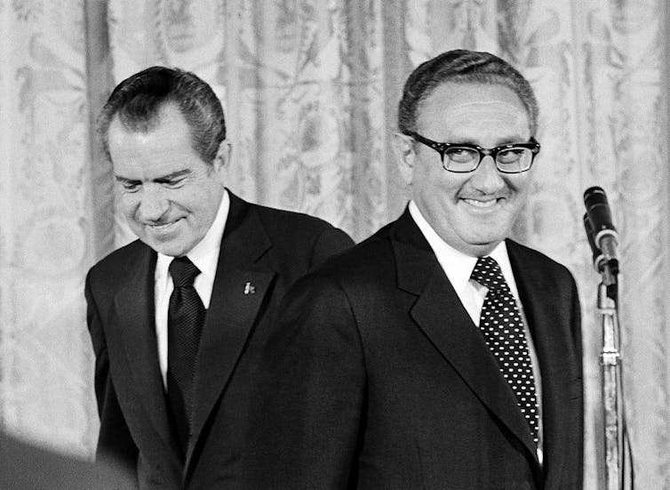 Henry Kissinger stands before a microphone in front of Richard Nixon.