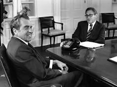 Nixon sits at his desk in the Oval Office smiling as Kissinger sits in front of the desk.