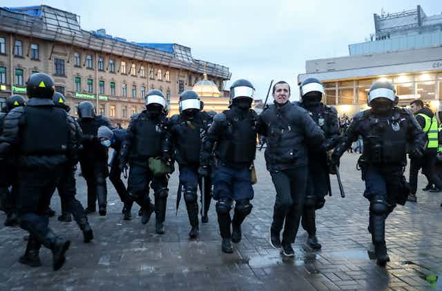 Police in riot gear walk a man in handcuffs through a public square