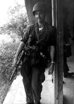 Daniel Ellsberg leans against a pole wearing army fatigues and holding a gun.