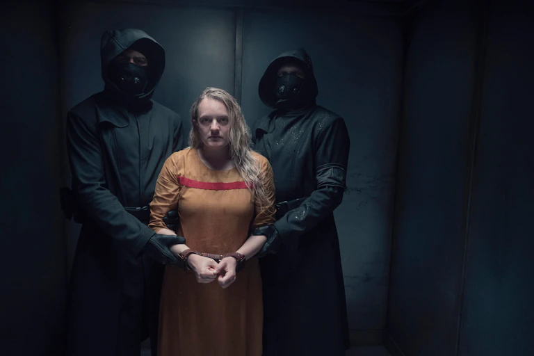 Production image: June held by two figures in black.