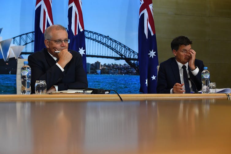 Scott Morrison and Angus Taylor sit with hands on their faces in front of Australian flags