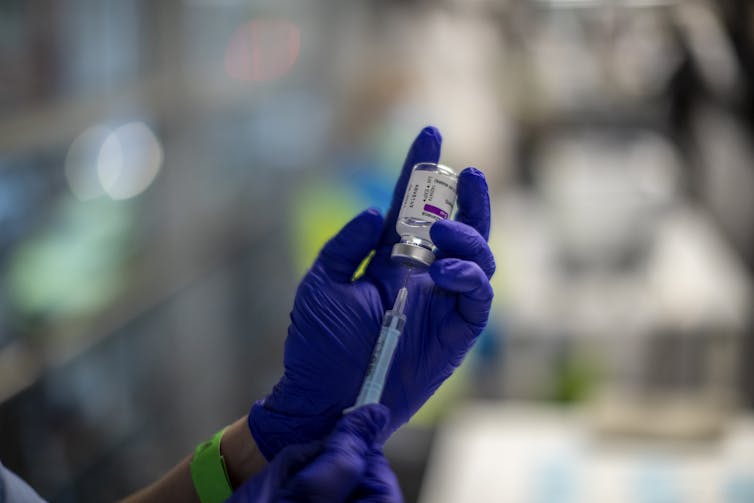 Gloved hands prepare a syringe from a vial of AstraZeneca vaccine.