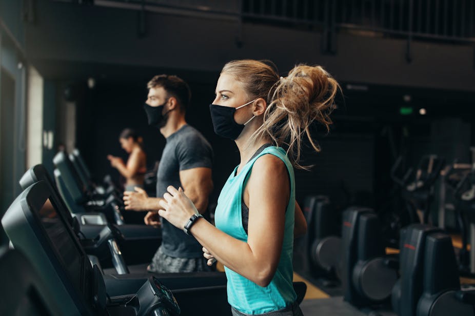 A man and a woman run on treadmills in the gym while wearing masks.