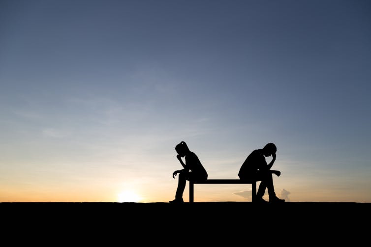 Two people sitting on a bench, with sunset in the background, with their heads in their hands.