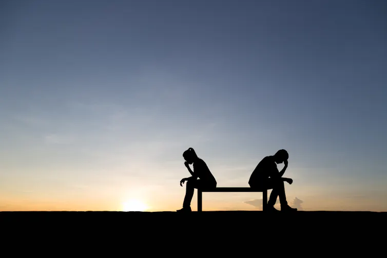 Two people sitting on a bench, with sunset in the background, with their heads in their hands.