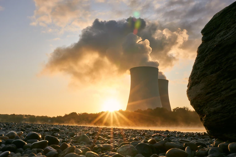 Two towers emit steam against a blue sky, with a low sun behind them. Pebbles can be seen in the foreground.