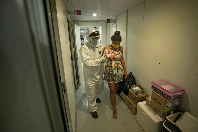 Profissional de saúde com EPI de corpo inteiro leva uma mulher grávida por um corredor