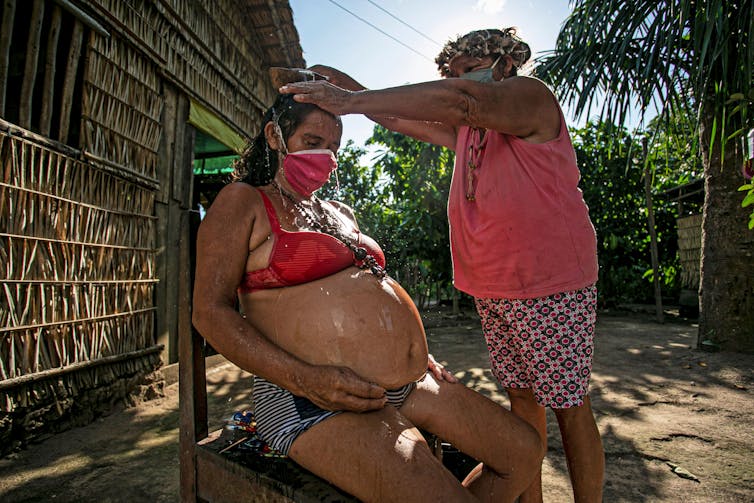 Uma mulher mais velha derrama água em uma mulher mais jovem usando uma máscara facial e biquíni com uma barriga de bebê muito grande