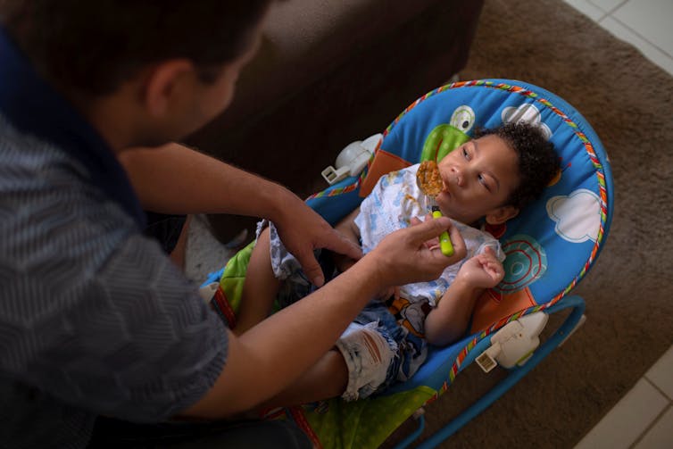 Man spoon feeds his toddler child, lying in a baby seat