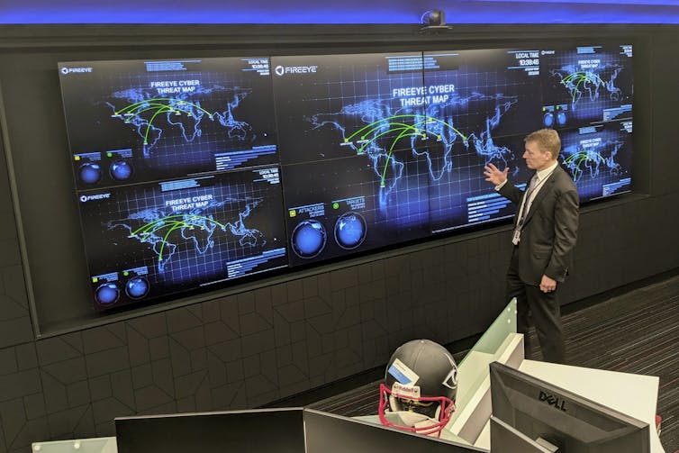 A man stands in front of a wall covered with computer displays showing maps of the world