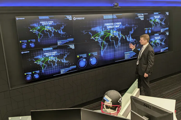 A man stands in front of a wall covered with computer displays showing maps of the world