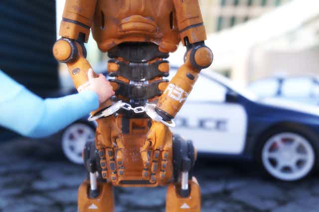 A humanoid robot stands with its arms in handcuffs behind its back, a blue-sleeved human arm holds one of the robot's arms, a police car in the background