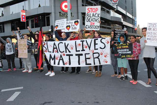 A group of people stand on a main road with banners that read 'Black lives matter' 'stop black deaths in custody and 'justice for fella''