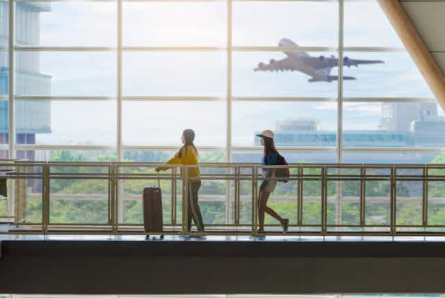 Two students walk through an airport terminal with a plane taking off outside