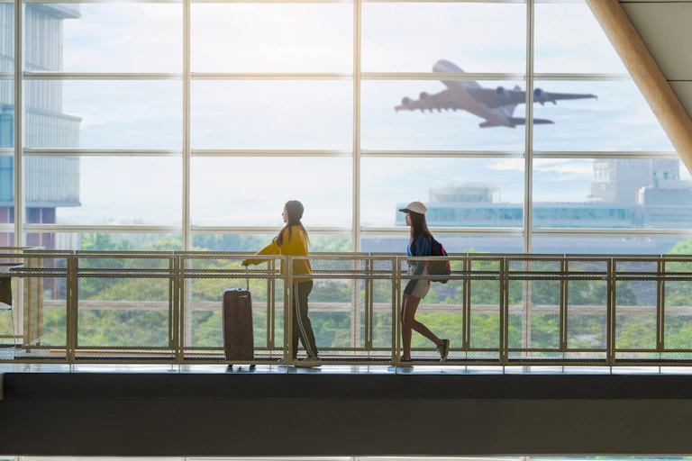 Two students walk through an airport terminal with a plane taking off outside