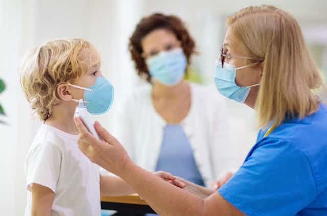 Una sanitaria toma la temperatura a un niño en presencia de otra mujer, los tres con mascarillas..