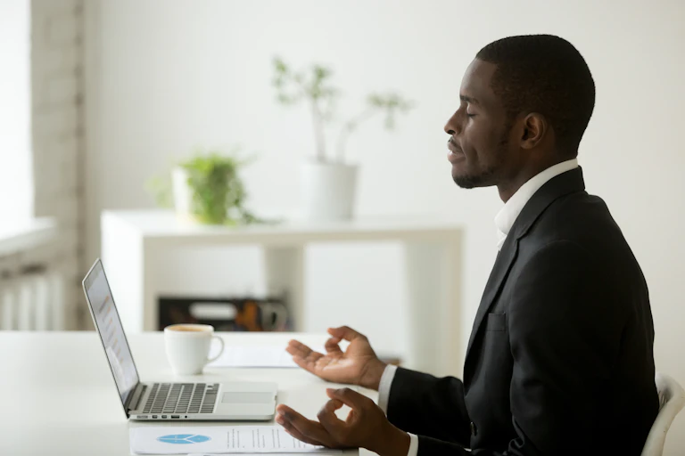 Man in suit sitting at a desk in front of a laptop, his eyes closed, index fingers and thumbs touching