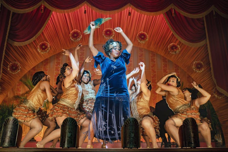 Black woman in blue dress dances on stage with backup dancers