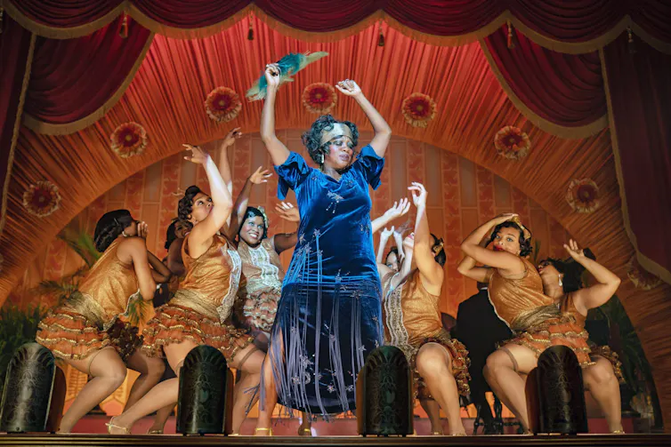 Black woman in blue dress dances on stage with backup dancers