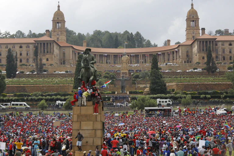 Crowd of people on the lawns outside large building, with statues visible