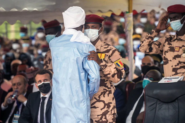 A man in a maroon military fatique and beret and Covid mask hungs another wearing a long-flowing garment and a turban
