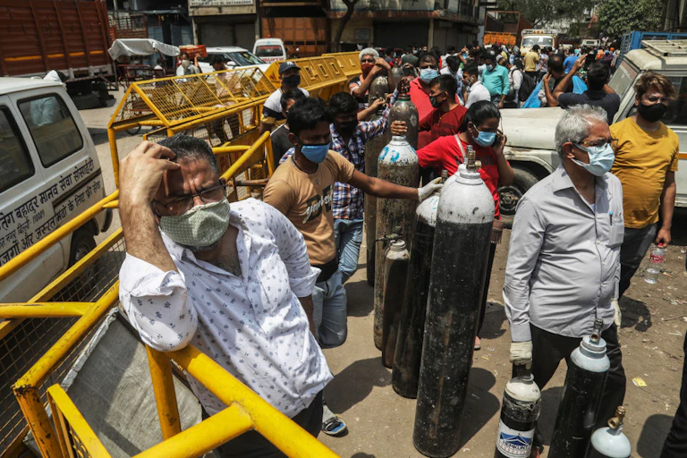 People wearing face masks stand in a line along a road, next to oxygen cylinders