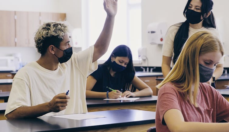 High school students and their teacher both wear masks in a classroom.