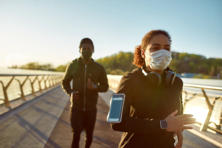 Una mujer y un hombre con mascarillas y ropa deportiva corren por la pasarela de una playa.