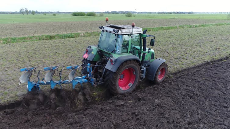 A green tractor uses a trailer to till the soil behind it.