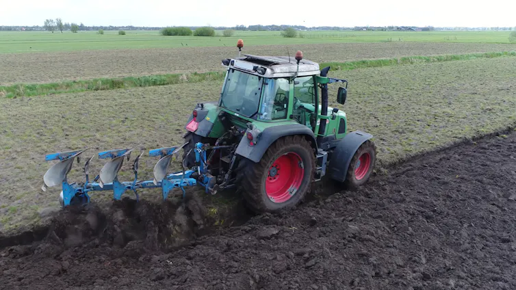 A green tractor uses a trailer to till the soil behind it.