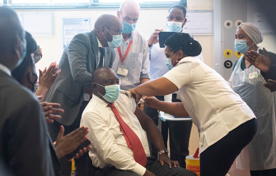 A seated man wearing a mask, with one shirt sleeve rolled up, receives an injection from a woman in nurse's uniform while other masked people look on