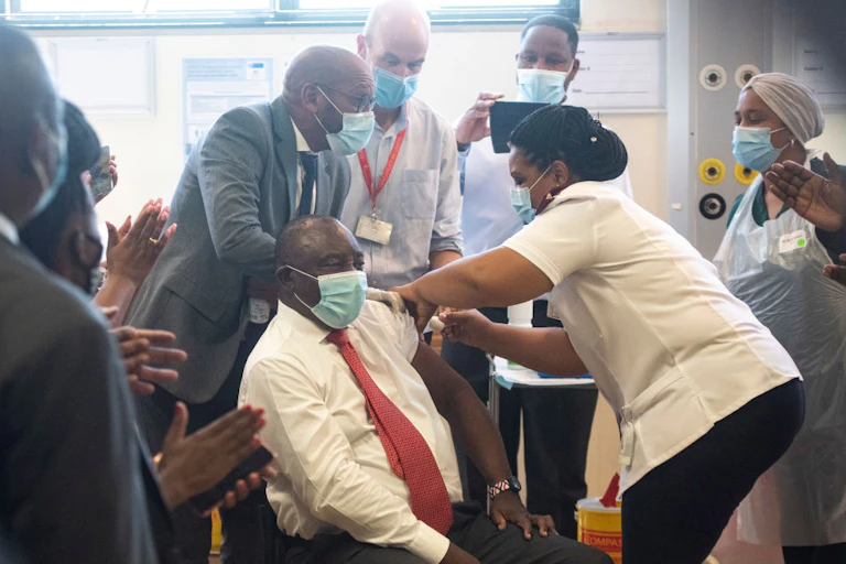 A seated man wearing a mask, with one shirt sleeve rolled up, receives an injection from a woman in nurse's uniform while other masked people look on