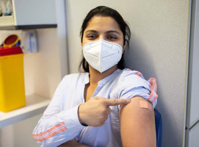 masked woman points to her vaccine bandaid
