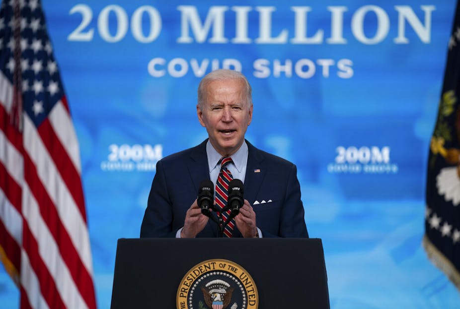 President Joe Biden speaks from a lecturn decorated with the US presidential seal. Behind him a backdrop reads: 200 million covid shots.