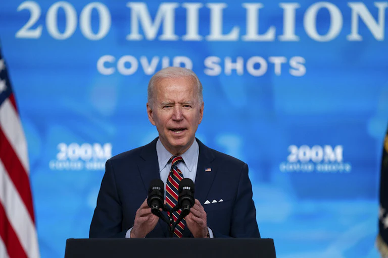 President Joe Biden speaks from a lecturn decorated with the US presidential seal. Behind him a backdrop reads: 200 million covid shots.