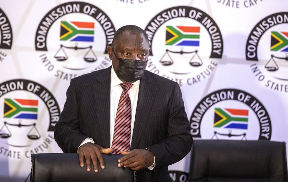 A man wearing a dark suit, white shirt, striped maroon tie and a black face mask stands behind a chair with the emblem of the South Africa's state capture commission in the background