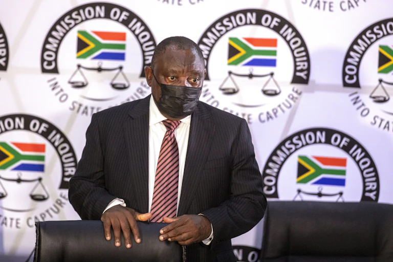 A man wearing a dark suit, white shirt, striped maroon tie and a black face mask stands behind a chair with the emblem of the South Africa's state capture commission in the background